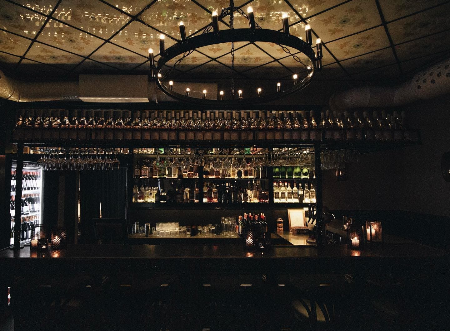 Almazas bar i levande ljus under en järnkrona, med flaskor och glas på hyllorna bakom disken